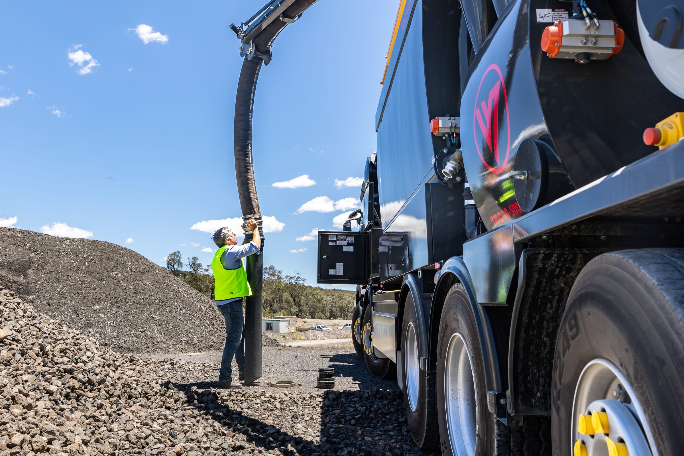 An operator in hi-vis guides the truck's large suction hose down into a pile of gravel.