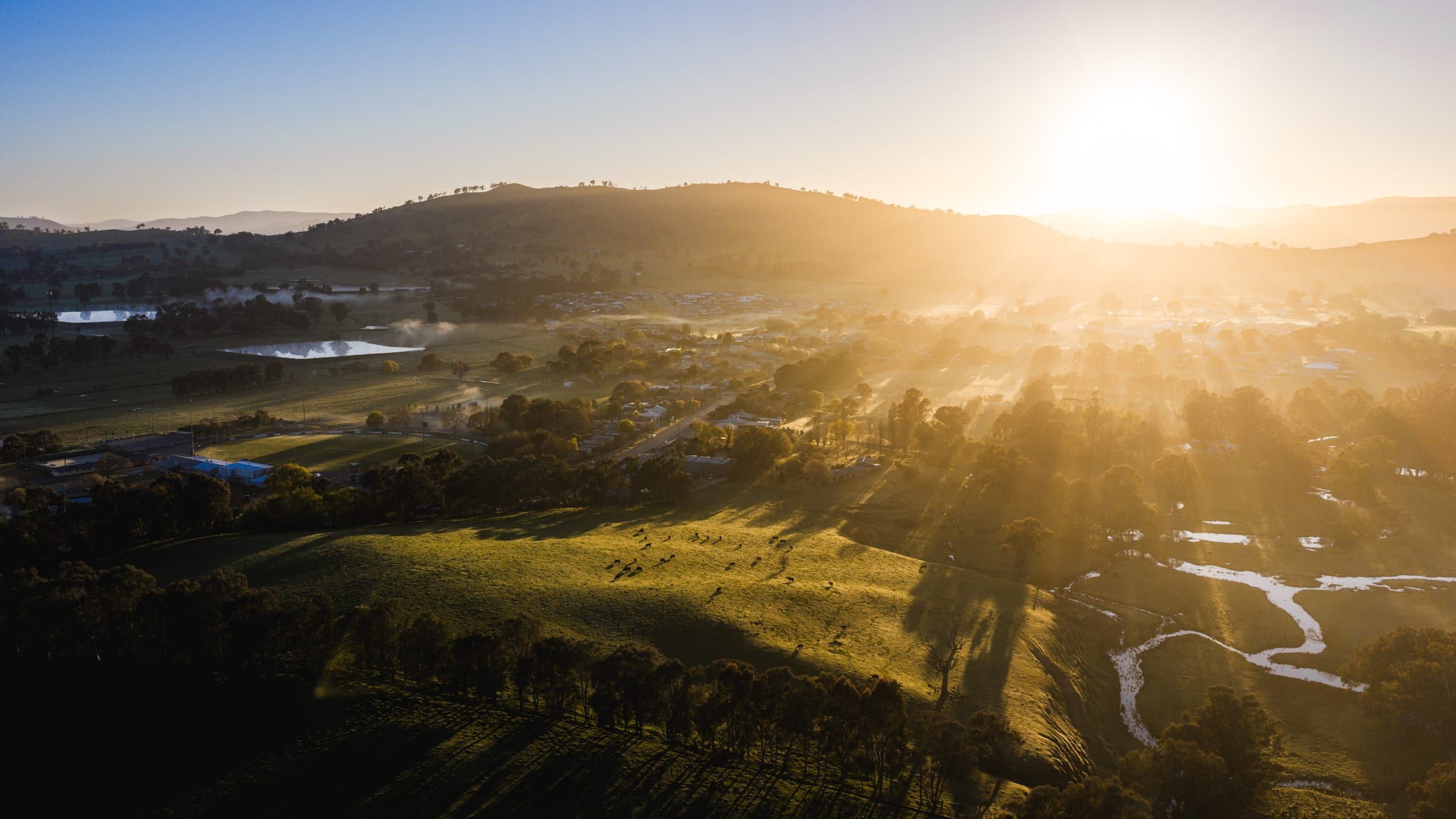 The sun rising over a ridge, casting long shadows across paddocks and a winding creek.