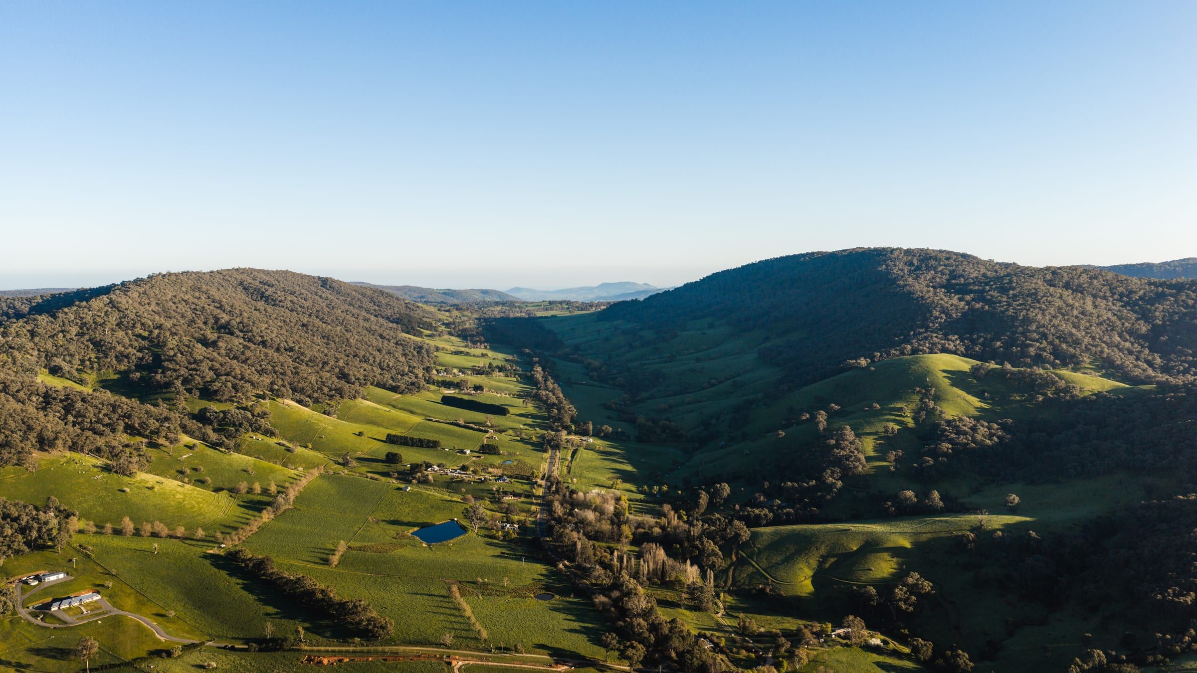 Rolling green hills and forested slopes, with a small dam in the paddocks below.