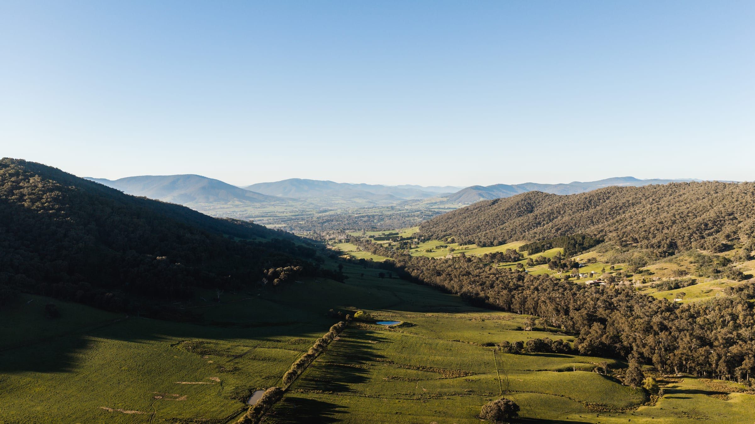 A shaded valley opening onto sunlit paddocks, with mountain ranges in the distance.