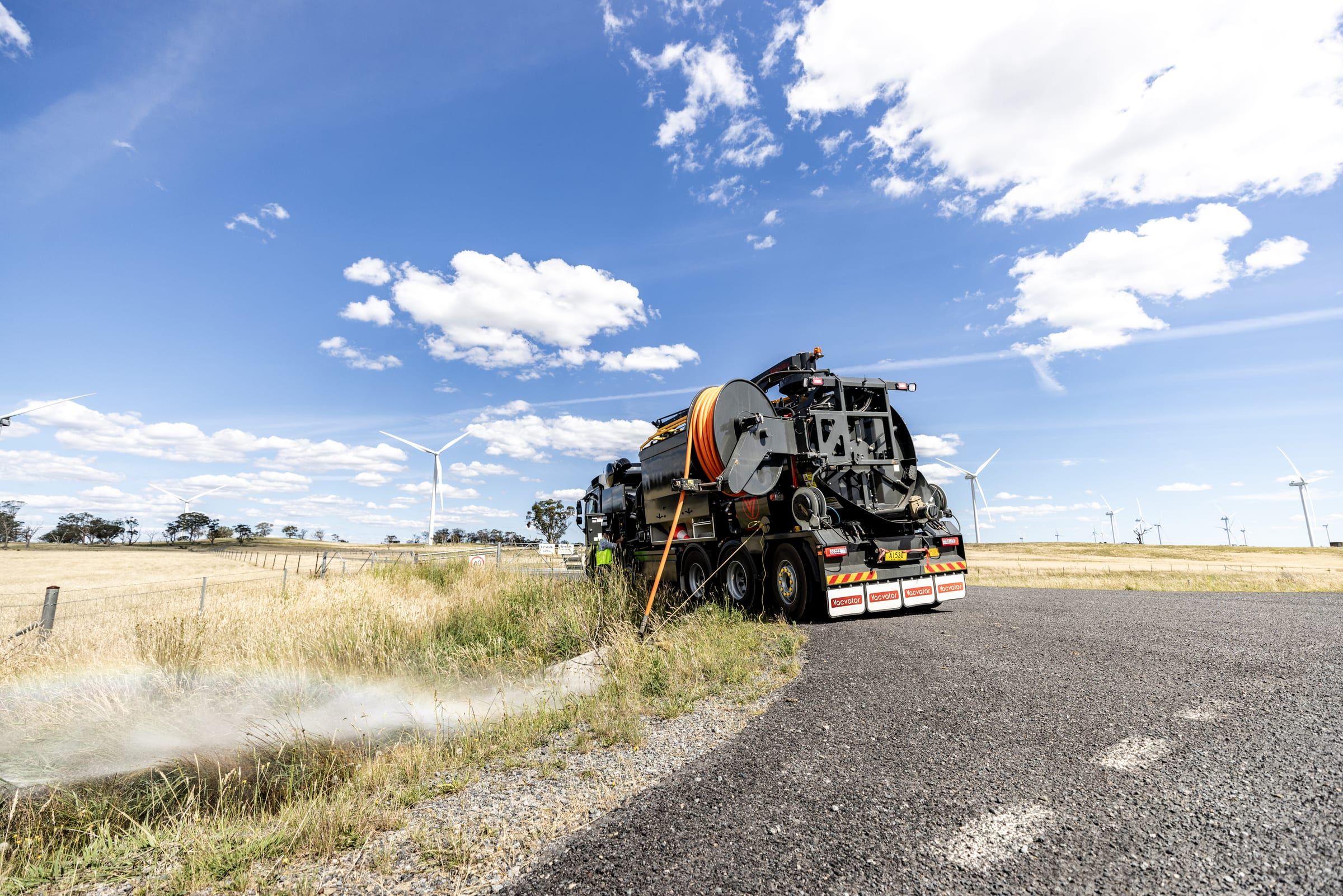 The truck parked by a country road, spray rising from its hose, with wind turbines on the hills behind.