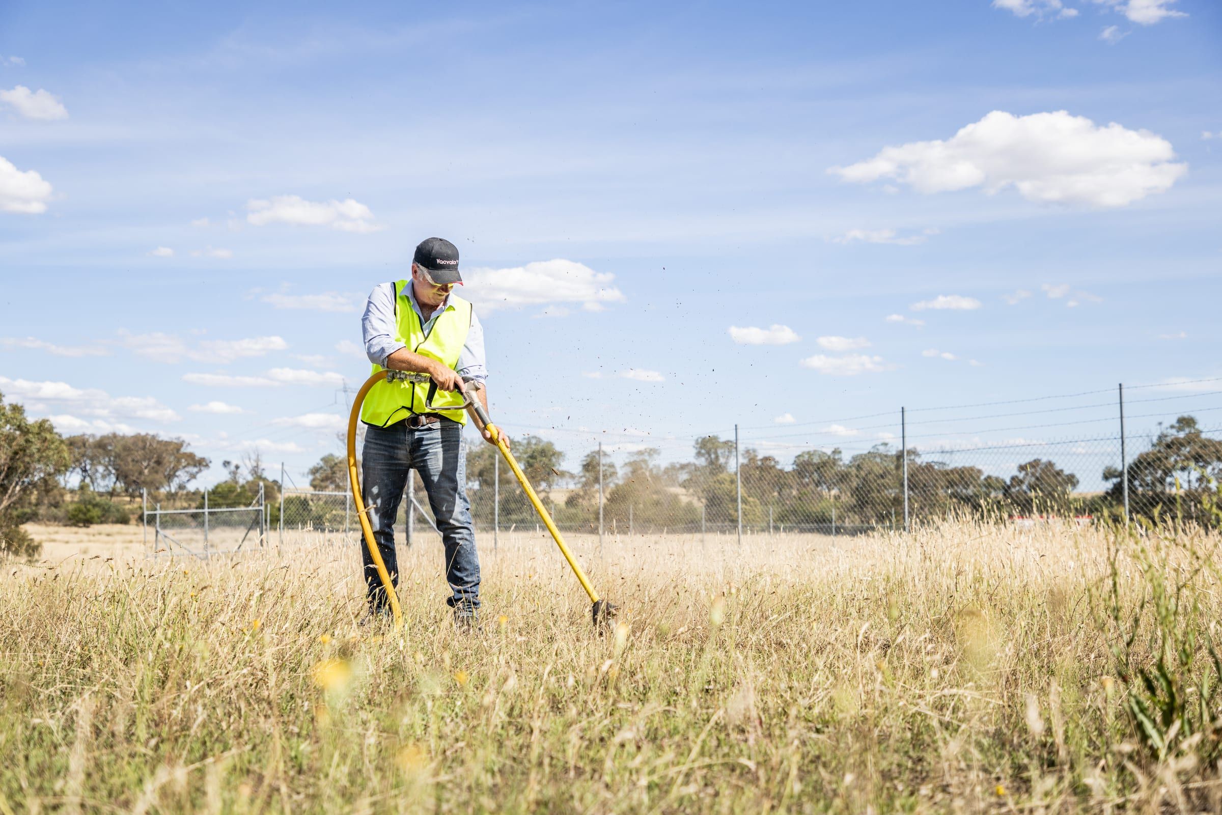 An operator in hi-vis working a digging lance in long dry grass, a hose trailing behind.