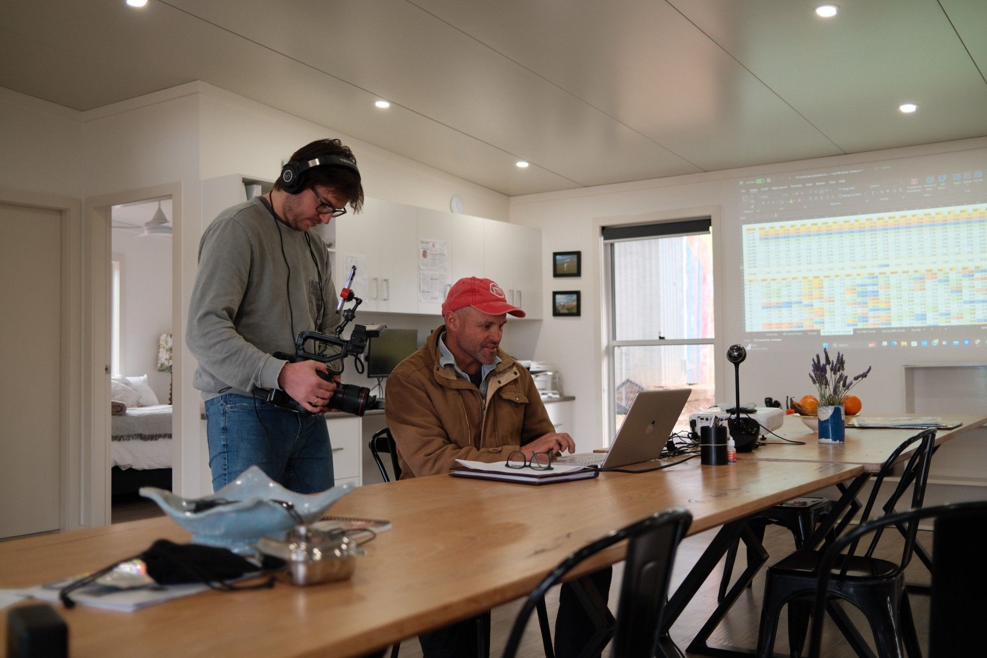 A camera operator in headphones films a man in a red cap working on a laptop, with a spreadsheet projected on the wall behind them.