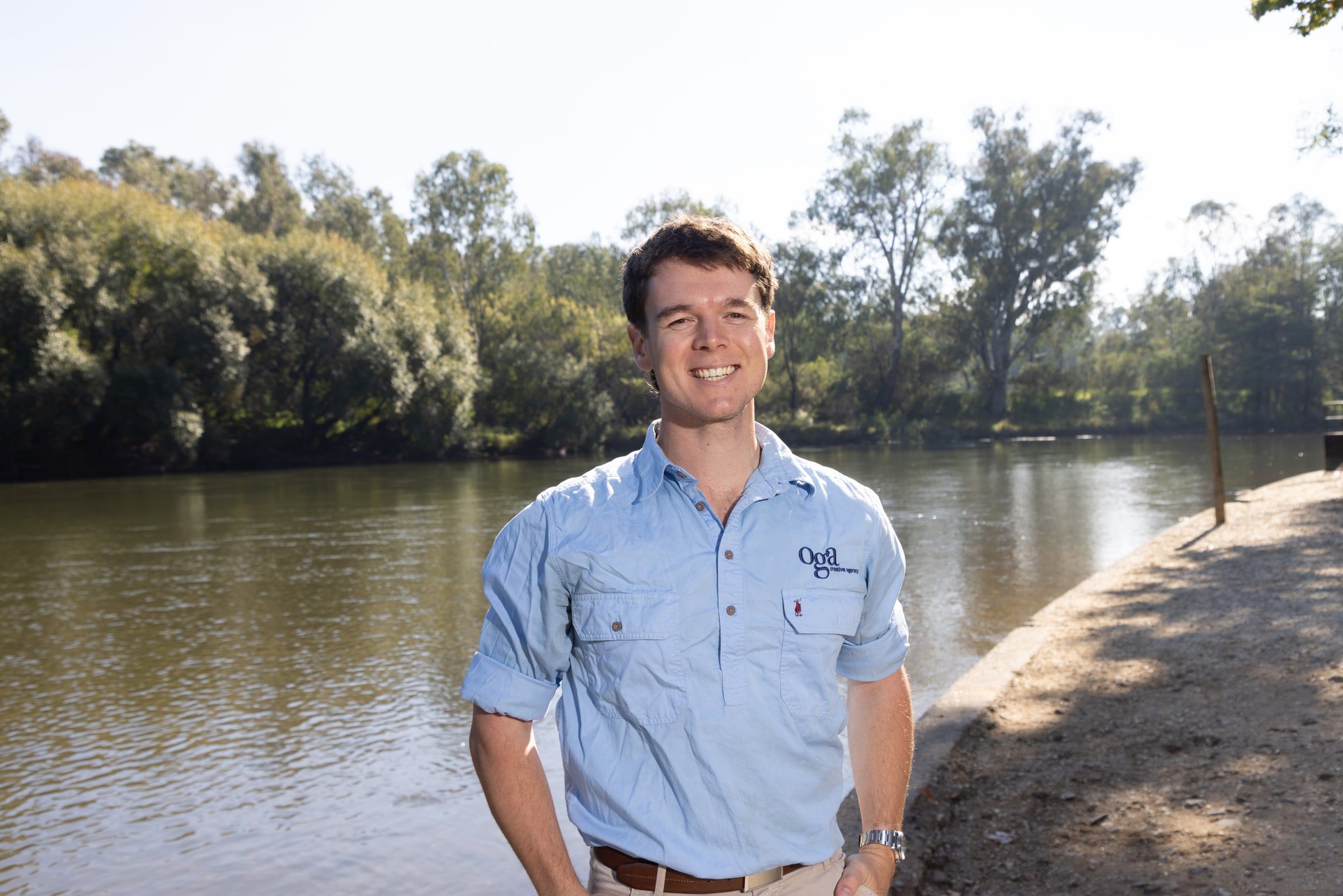 Zac Edmonds smiling on a sunny riverbank lined with gum trees, wearing a light blue Oga Creative work shirt.