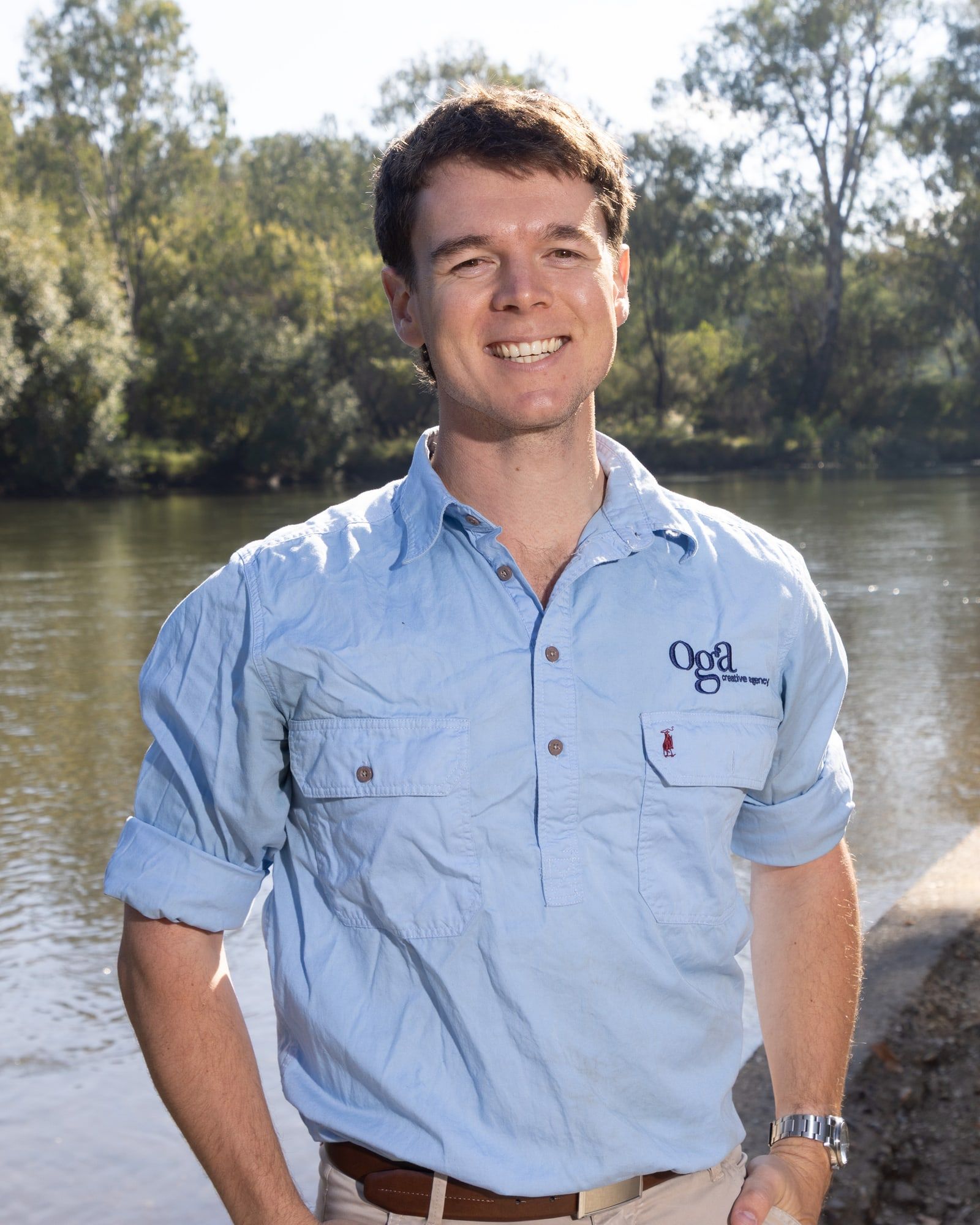 Zac Edmonds smiling on a sunny riverbank lined with gum trees, wearing a light blue Oga Creative work shirt.
